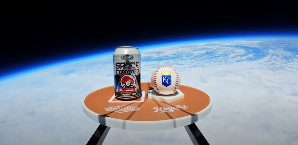 A can of Space Camper Cosmic IPA and a Kansas City Royals baseball sit on a small round table with "Boulevard" and "Royals" logos, floating high above Earth with the curvature and atmosphere visible in the background.